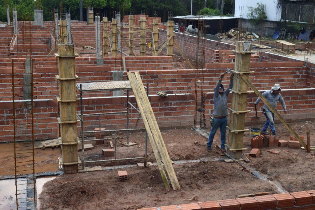 trabalhadores realizam serviços de alvenaria e montagem de formas para concreto entre os ambientes da construção.