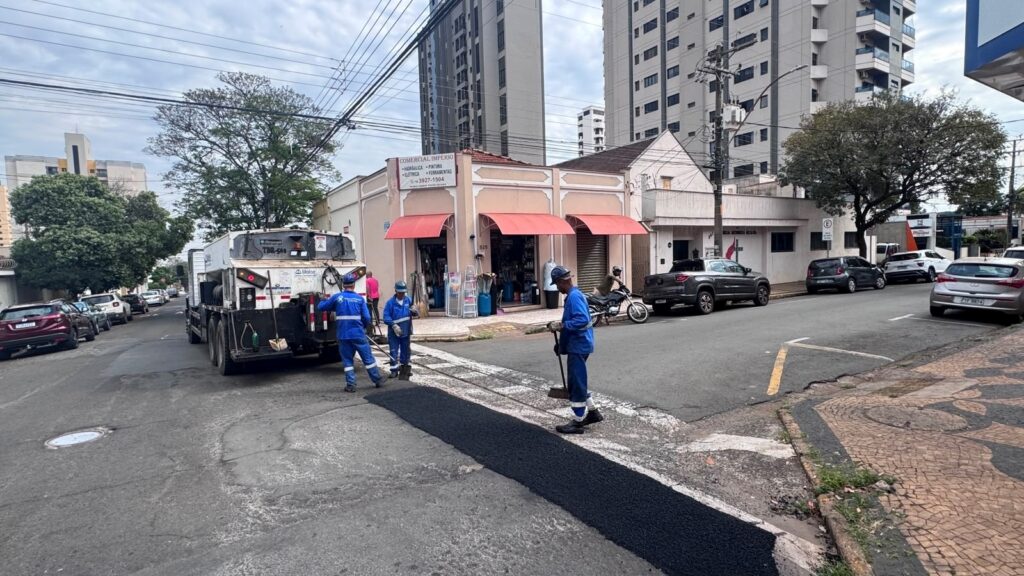 A imagem mostra uma rua urbana movimentada, com três trabalhadores usando uniformes azuis fazendo reparos no asfalto em uma faixa de pedestres. Ao lado deles há um caminhão de manutenção. No centro, aparece uma loja de materiais para construção chamada “Comercial Império”, com toldos vermelhos e produtos expostos na calçada. A rua tem vários carros estacionados, uma motocicleta, árvores, muitos fios elétricos e prédios residenciais altos ao fundo. O céu está nublado e as calçadas têm o tradicional padrão de pedras portuguesas.