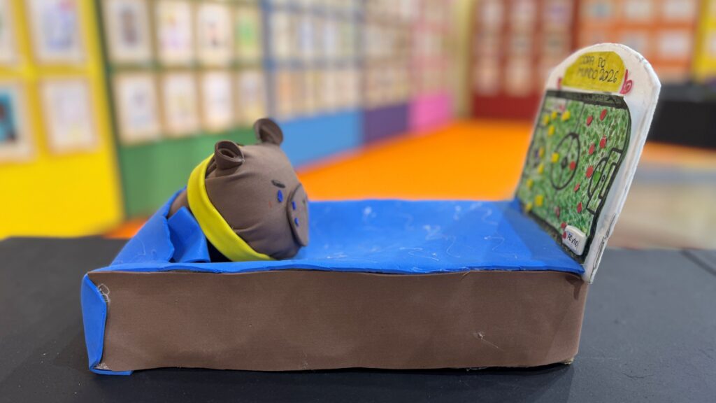 A imagem mostra uma maquete artesanal feita com materiais escolares. Em primeiro plano, há um personagem marrom deitado em uma cama azul, com a cabeça apoiada em uma espécie de travesseiro amarelo.