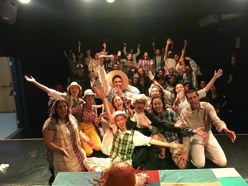 A imagem mostra um grande grupo de pessoas reunidas em um palco de teatro, posando para uma foto coletiva