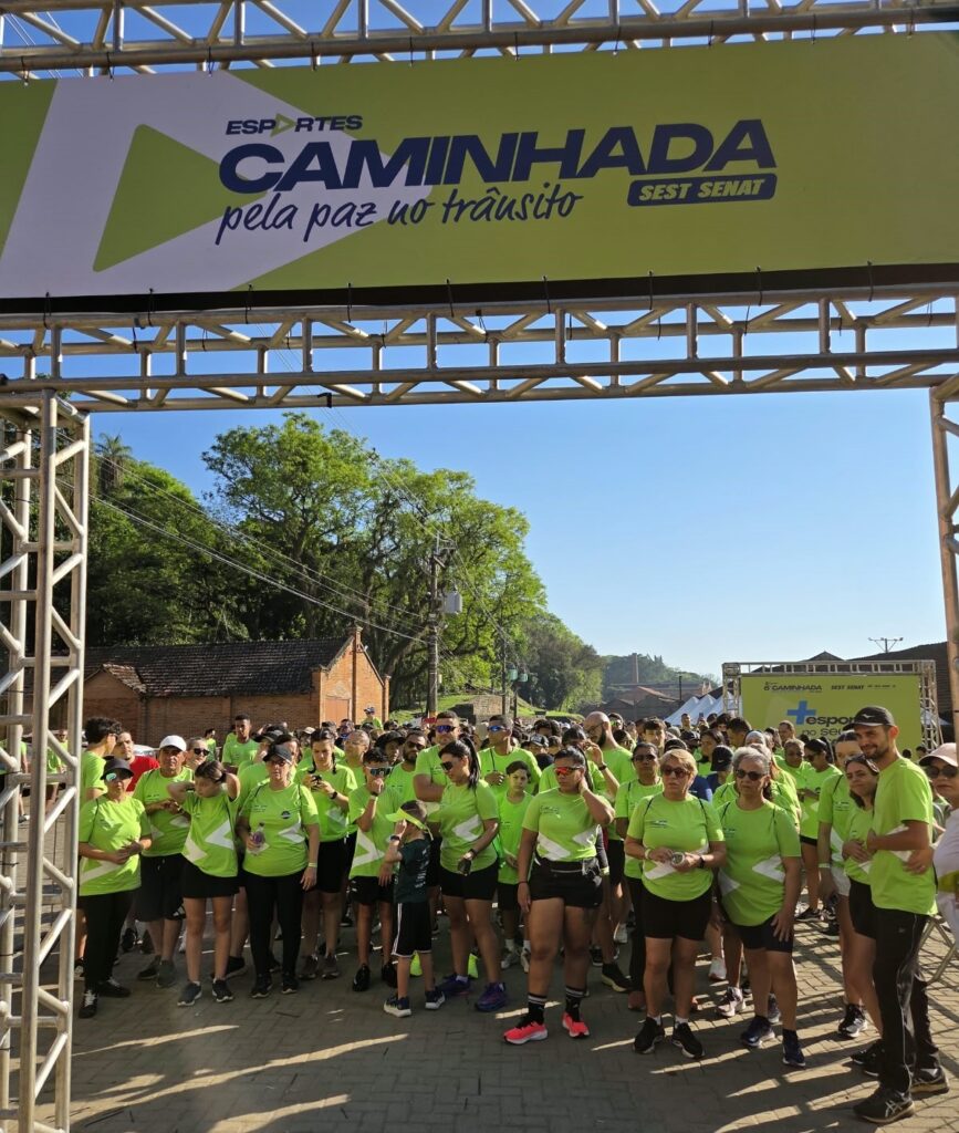 Um grupo numeroso de participantes, vestindo camisas verdes combinadas, reúne-se na linha de partida sob um pórtico metálico com a faixa "Caminhada pela paz no trânsito SEST SENAT".