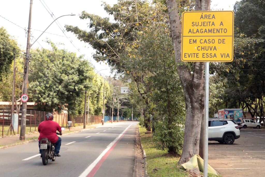 A imagem mostra uma via urbana arborizada, com um motociclista trafegando e uma placa de alerta em destaque. A placa amarela informa: “Área sujeita a alagamento. Em caso de chuva, evite essa via.”