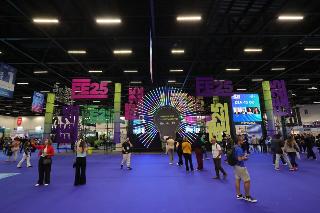 Vista geral de uma feira de negócios e inovação, com visitantes circulando por uma área temática identificada como “Arena do Conhecimento” e estruturas decoradas com a marca do evento.