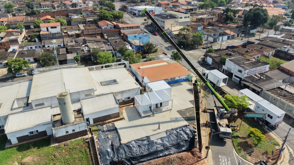 Vista aérea mostra a instalação de módulos pré-fabricados em unidade de saúde, com guindaste e estrutura em processo de ampliação.