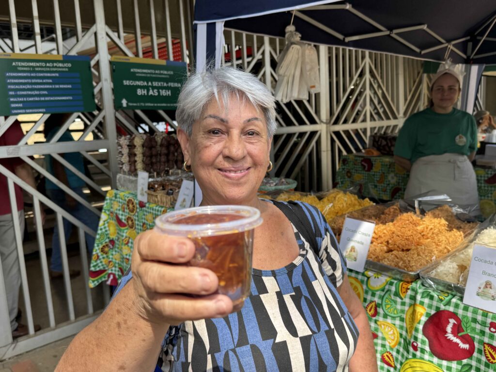 A imagem mostra uma mulher idosa segurando um recipiente bandeja transparente com doces. Ao fundo, há uma grade metálica e uma placa verde com informações sobre serviços públicos, incluindo “Atendimento ao público”, “Segunda a sexta-feira” e “6h às 18h”.