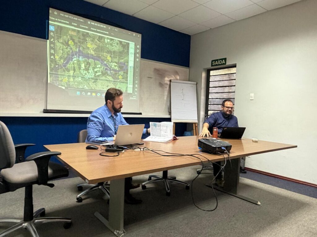 A imagem mostra uma reunião técnica em uma sala de conferências. Dois homens estão sentados à mesa, cada um com um notebook, enquanto um projetor exibe na tela um mapa geográfico com marcações e coordenadas — possivelmente usado para análise territorial ou planejamento estratégico. Há também um quadro branco e um flip chart ao fundo, reforçando o ambiente profissional e de discussão técnica.