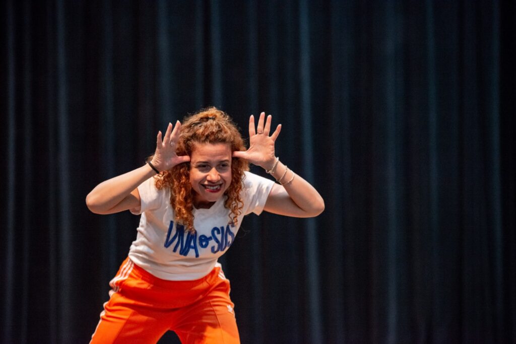 A imagem mostra uma pessoa em um palco com fundo escuro, usando camiseta branca e calça laranja