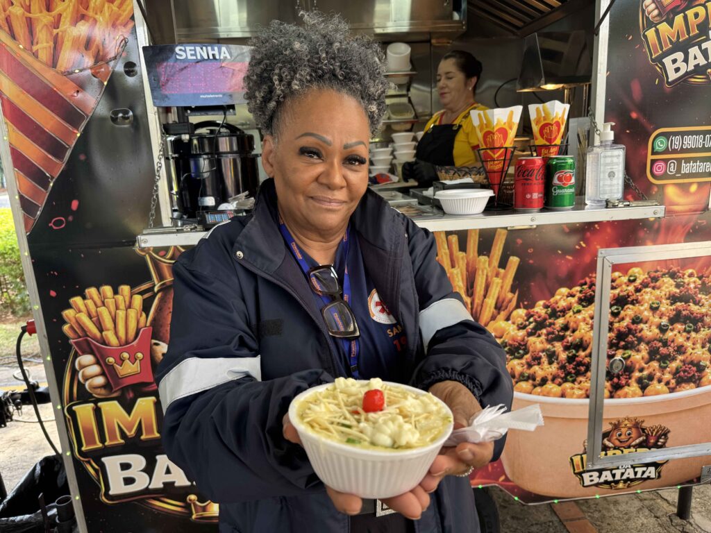 A foto mostra uma mulher de cabelos grisalhos, com óculos escuros pendurado e uniforme azul-marinho. Ela está atrás de um balcão de lanchonete, segurando um prato com batatas fritas cremosas. Ao fundo, há um freezer ou expositor com garrafas de refrigerante, incluindo Coca-Cola e Guaraná Antarctica, além de embalagens de alimentos. O balcão tem um painel colorido com imagens de hambúrgueres e batatas.