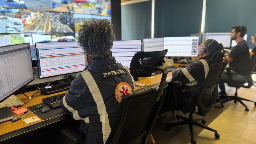A imagem mostra uma central de monitoramento e atendimento de emergências. Em primeiro plano, uma profissional usando uniforme com a identificação “ENFERMEIRA” trabalha diante de vários monitores. Outros funcionários, alguns usando fones de ouvido e uniformes com faixas refletivas, também operam computadores. As telas exibem sistemas de atendimento e informações operacionais, enquanto um grande painel ao fundo mostra imagens de câmeras de trânsito e vias urbanas.