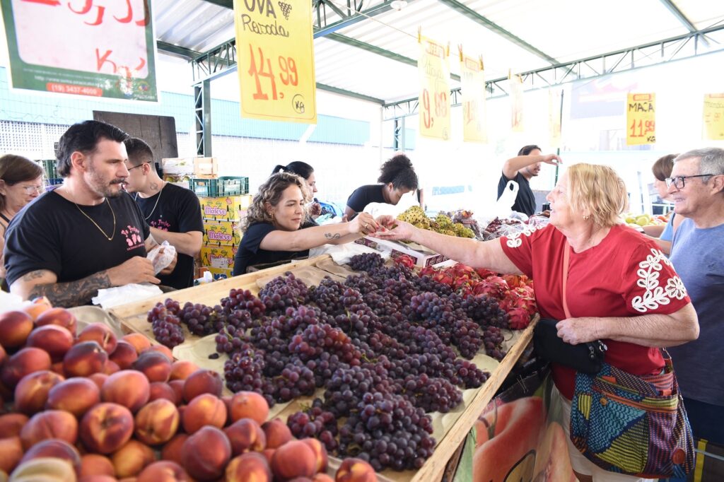 A imagem mostra uma feira de frutas em um espaço coberto. No primeiro plano, há grandes caixas com pêssegos, uvas roxas e pitaias. Várias pessoas estão comprando e atendendo na banca. À direita, uma senhora de blusa vermelha conversa com um homem de óculos, enquanto uma mulher ao centro entrega uma fruta a ela. Ao fundo, há placas de ofertas e preços, incluindo uva, mamão e outras frutas.