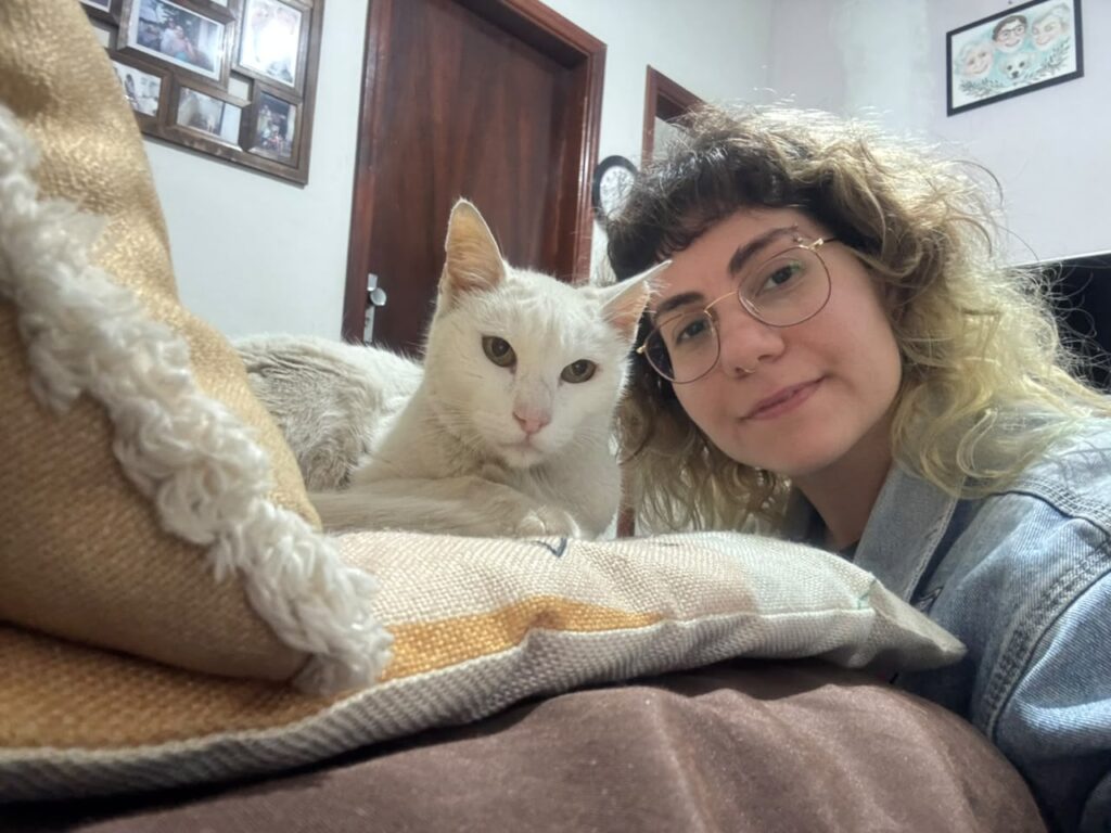 A imagem mostra uma jovem mulher tirando uma selfie fofa com seu gato branco em uma sala aconchegante. Na imagem, uma jovem mulher com óculos redondos e cabelo loiro ondulado está tirando uma selfie. Ela veste uma jaqueta jeans. Ao lado dela, um gato branco está deitado em uma almofada. O gato tem um olhar calmo e direto para a câmera. Atrás deles, um sofá marrom e almofadas decorativas. Ao fundo, há uma porta de madeira, quadros de fotos em uma parede e um pequeno relógio. A iluminação sugere que a foto foi tirada em um ambiente interno, possivelmente à noite ou com pouca luz natural. A atmosfera geral da imagem é relaxada, íntima e carinhosa, transmitindo um sentimento de companheirismo entre a mulher e seu animal de estimação