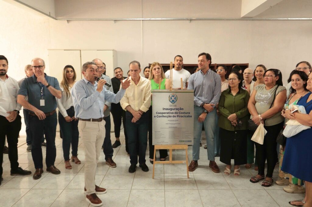 A imagem mostra uma cerimônia de inauguração em um espaço interno. Um homem segura um microfone e parece fazer um discurso, enquanto várias pessoas estão reunidas ao redor, posando para a foto. No centro, há um painel em um cavalete com o brasão de Piracicaba e o texto: “Inauguração Cooperativa de Costura e Confecção de Piracicaba”. O ambiente tem piso claro, paredes brancas e armários ao fundo.