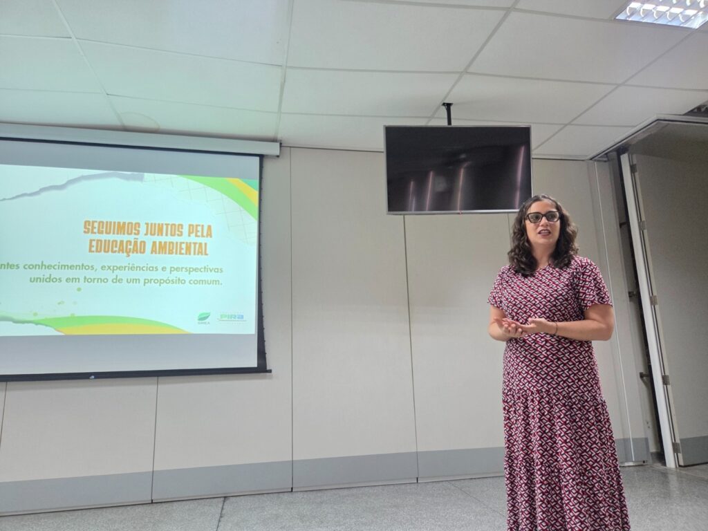A imagem mostra uma mulher apresentando em uma sala de reuniões. Ela usa óculos e um vestido estampado em vermelho, branco e preto, e está de pé ao lado de uma tela de projeção. No slide aparece a frase “Seguimos juntos pela educação ambiental”, acompanhada de um texto sobre conhecimentos, experiências e perspectivas unidos em torno de um propósito comum. Também aparecem logotipos relacionados à educação ambiental e à cidade de Piracicaba. Ao fundo, há uma televisão desligada, paredes claras, teto com placas e uma porta aberta.