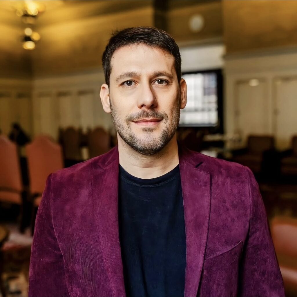 A imagem mostra um homem posando para um retrato em ambiente interno. Ele tem cabelo curto escuro, barba rala e expressão serena, olhando diretamente para a câmera. Veste um blazer vinho sobre uma camiseta azul-marinho. Ao fundo, há uma sala com iluminação quente, cadeiras, mesas e uma janela, tudo levemente desfocado.