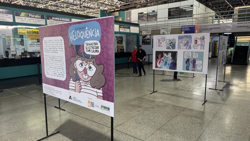 A imagem mostra uma exposição de histórias em quadrinhos montada dentro de um terminal rodoviário. Vários painéis estão organizados em fileiras, apoiados em estruturas pretas, com ilustrações coloridas, personagens e balões de fala. No centro, há um painel biográfico sobre a jornalista, ilustradora e quadrinista Helô D’Angelo. À direita, um grande quadrinho aborda situações do cotidiano e relacionamentos, com a frase em destaque “Trincas para se estressar com calma”. Ao fundo, aparecem placas do terminal indicando destinos como Goiânia, Uberlândia, Curitiba, Florianópolis e Ouro e Prata, além de áreas de embarque, despacho de encomendas e banheiros.