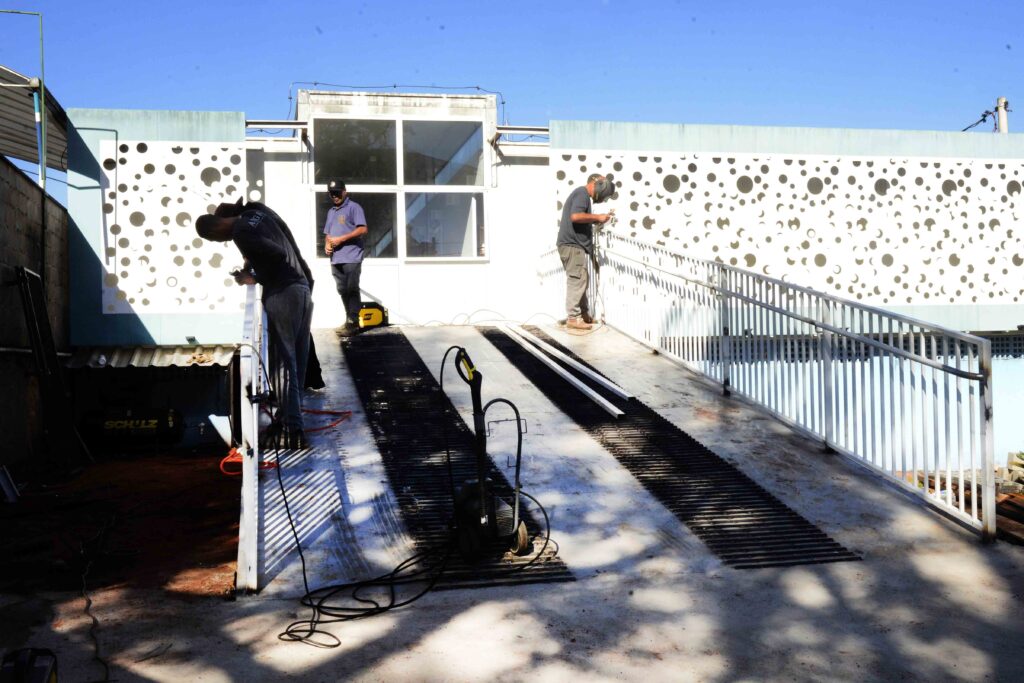 A imagem mostra uma equipe trabalhando na instalação ou manutenção de um sistema de energia solar no telhado de um prédio. Duas pessoas aparecem sobre a cobertura: uma usa uniforme azul e segura uma ferramenta, enquanto a outra veste roupas escuras e parece manusear um painel ou equipamento. Há vários cabos espalhados pelo chão, estruturas metálicas, placas solares parcialmente visíveis e uma ferramenta elétrica amarela. O local tem grades de proteção, paredes brancas e azuis e uma forte incidência de luz solar. A imagem apresenta muitas manchas circulares e áreas superexpostas, que dificultam a visualização de alguns detalhes.
