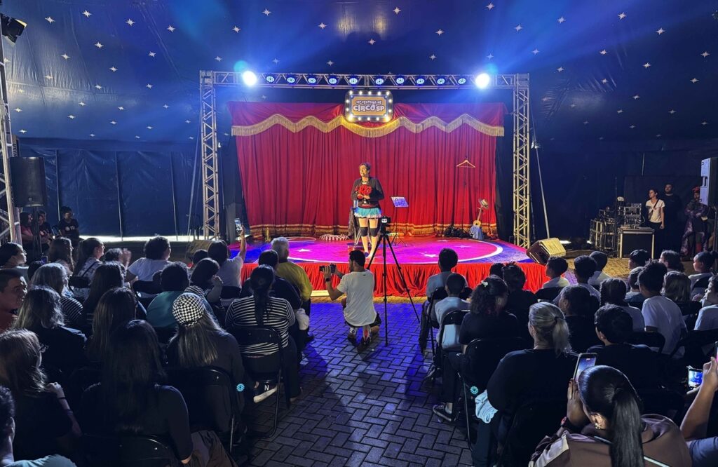 A imagem mostra uma apresentação circense em uma tenda iluminada de azul, decorada com pequenas estrelas no teto. No centro do palco, uma artista vestida como palhaça segura flores diante de uma cortina vermelha com detalhes dourados. Uma placa acima do palco anuncia o 15º Festival de Circo SP. O público está sentado em cadeiras, assistindo atentamente; algumas pessoas gravam a apresentação com celulares. Há equipamentos de som e iluminação nas laterais, além de outros artistas ou integrantes da equipe próximos ao palco.
