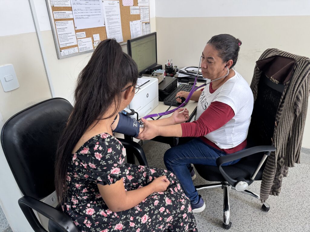 A imagem mostra uma profissional de saúde realizando a medição de pressão arterial de uma paciente em um consultório. A profissional usa um estetoscópio e um esfigmomanômetro, enquanto a paciente, vestida com um vestido floral, está sentada e colaborando com o procedimento.