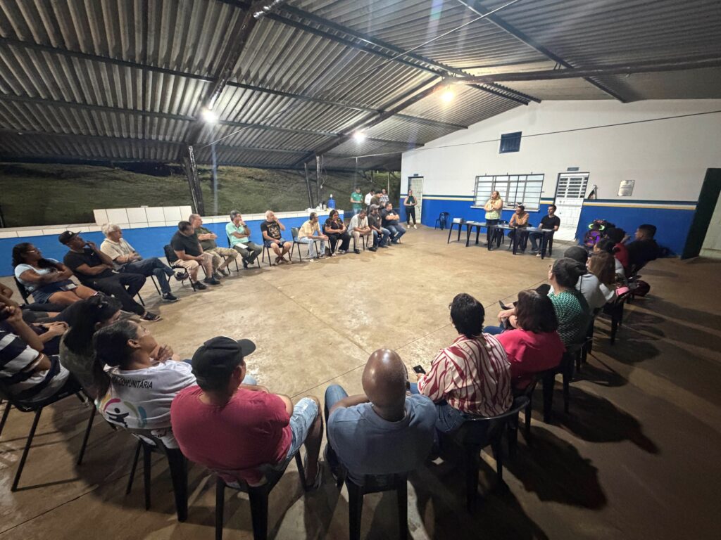Reunião comunitária realizada em um espaço coberto, com dezenas de pessoas sentadas em círculo. Ao fundo, há uma mesa com participantes conduzindo a conversa ou apresentação.