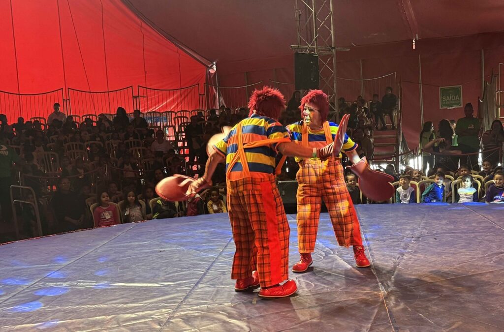 A imagem mostra dois palhaços se apresentando no picadeiro de um circo. Eles usam perucas vermelhas, maquiagem colorida, camisetas listradas em azul, amarelo e laranja, suspensórios laranja e calças xadrez vermelhas. Um deles está de costas, segurando uma grande luva ou objeto vermelho, enquanto o outro faz um gesto teatral com as mãos. Ao fundo, há uma plateia sentada em cadeiras plásticas, incluindo várias crianças. A tenda é vermelha, com equipamentos de som e iluminação, além de uma placa indicando a saída de emergência.