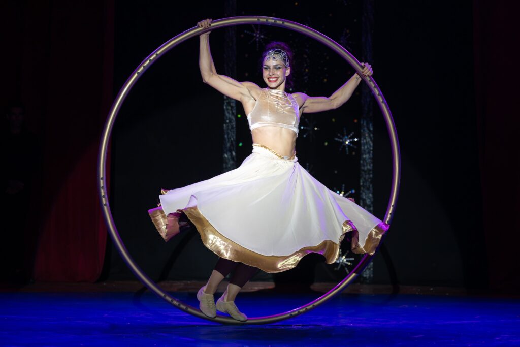 A imagem mostra uma artista circense realizando uma performance com um Cyr wheel — um grande aro metálico usado em acrobacias e dança. Ela está equilibrada dentro do aro, com os braços estendidos e um sorriso confiante, vestindo uma roupa elegante de top claro e saia branca com detalhes dourados. O fundo escuro e as luzes destacam sua figura, criando uma atmosfera de espetáculo e graça.
