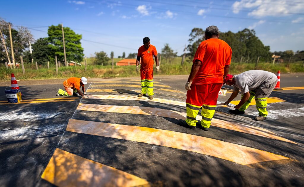 A imagem mostra trabalhadores realizando a pintura de sinalização em uma estrada. Eles usam uniformes laranja e calças com faixas refletivas amarelas; alguns estão ajoelhados aplicando tinta, enquanto outros observam ou trabalham ao redor. O asfalto está coberto por faixas e áreas de tinta amarela e branca, com cones de trânsito e latas de tinta espalhados pelo local. Ao fundo há uma paisagem rural, com árvores, cercas, vegetação e céu azul com nuvens.