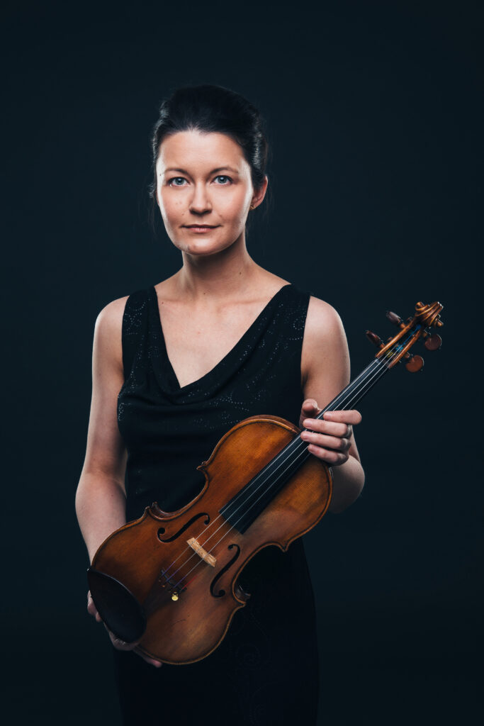 Mulher posa para um retrato, usando um vestido preto e segurando um violino, em frente a um fundo escuro.
