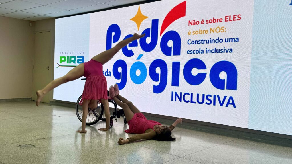 A imagem mostra duas bailarinas realizando uma apresentação artística em um ambiente interno, diante de um grande painel digital com o título “Pedagógica Inclusiva” e o logotipo da Prefeitura de Pira.