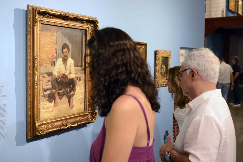 Visitantes observam atentamente uma pintura emoldurada em uma exposição de arte, dentro de uma galeria com paredes azuis.