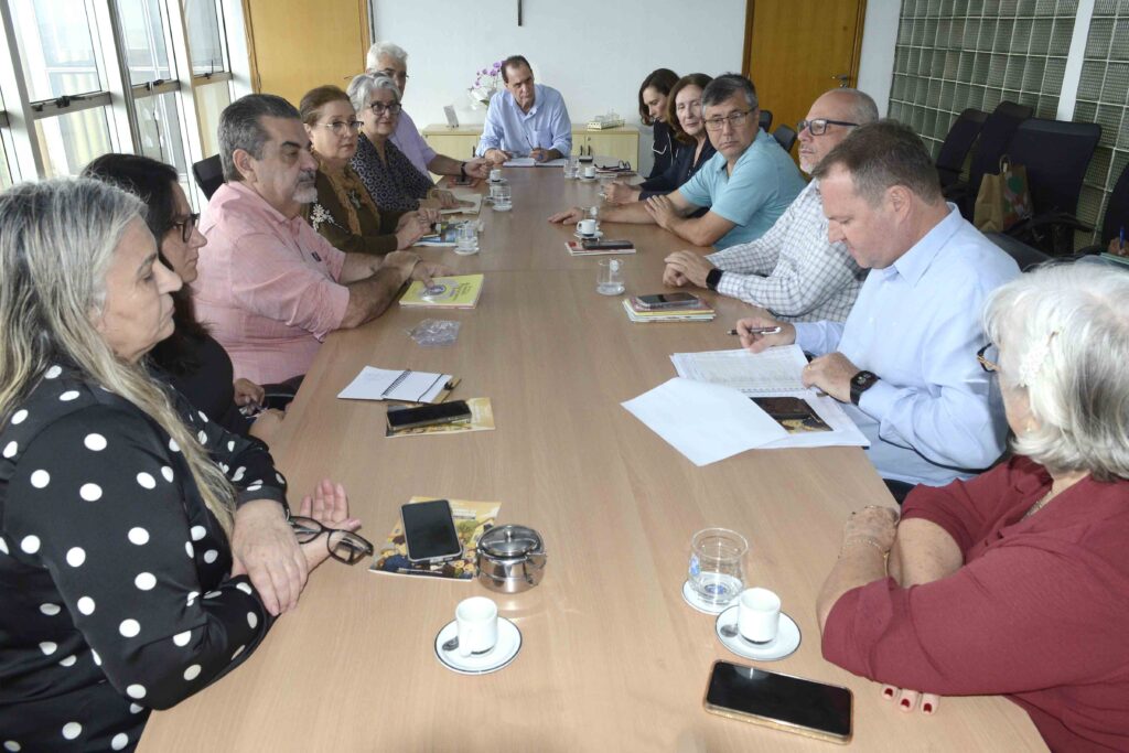 A imagem retrata outro momento de reunião formal, com várias pessoas sentadas em torno de uma longa mesa de conferência. Sobre a mesa estão espalhados papéis, cadernos, celulares, xícaras de café e copos de água, elementos que reforçam o caráter prático e cotidiano da discussão. Um dos participantes escreve em um documento, indicando que decisões ou registros estão sendo feitos em tempo real. O ambiente é iluminado por grandes janelas e possui detalhes como uma porta de madeira e uma parede com blocos de vidro, criando uma atmosfera profissional e colaborativa.