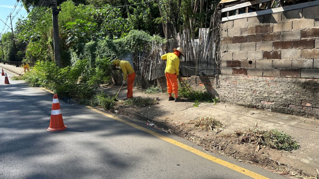 Trabalhadores com fardamento amarelo e taronja realizam serviços de capinação e corte de mato num passeio junto a um muro e a uma via sinalizada com cones de trânsito.