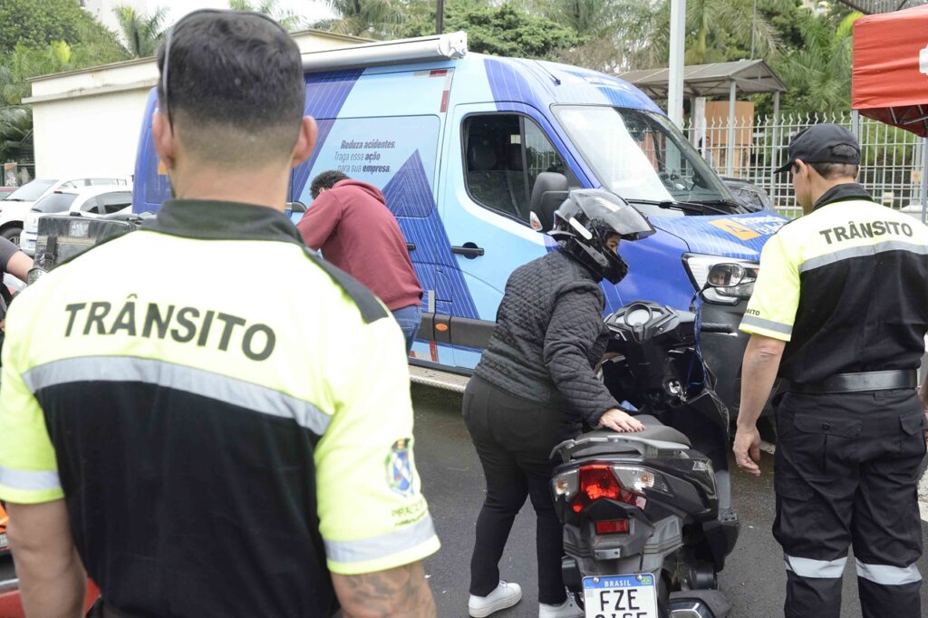 Agentes de trânsito devidamente uniformizados realizam uma abordagem educativa a uma condutora de scooter/moped em frente a uma carrinha de apoio do programa de trânsito.