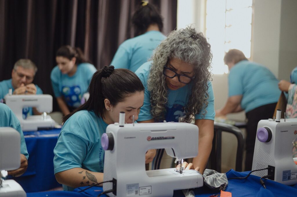 Uma atividade prática de costura, em que uma pessoa orienta outra no uso de uma máquina de costura. Ao fundo, outros participantes também trabalham e aprendem em um ambiente de capacitação.