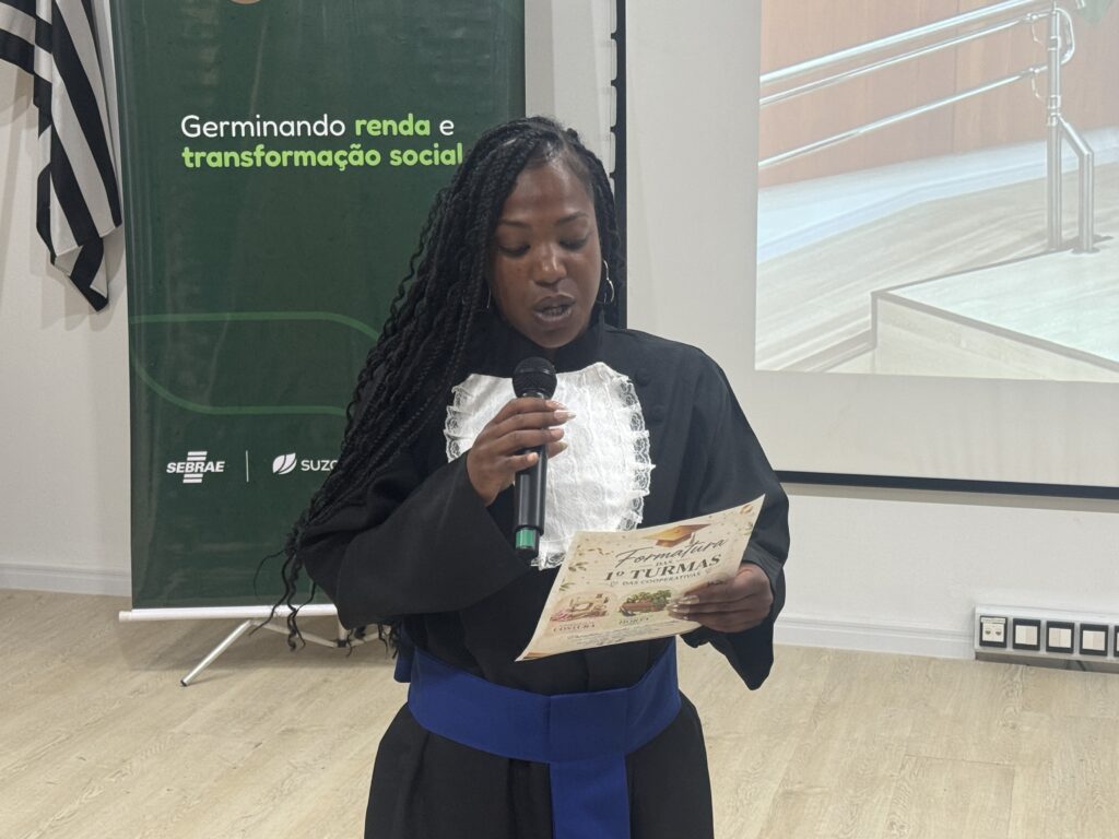 A imagem mostra uma mulher negra com longos cabelos trançados, usando brincos grandes e uma roupa preta com detalhes azuis. Ela segura um microfone e um folheto enquanto parece falar em um evento. Ao fundo, há um painel verde com os dizeres “Germinando renda e transformação social”, além dos logotipos do Sebrae e de outra instituição. À esquerda, aparece parcialmente uma bandeira listrada em preto e branco; à direita, há janelas com persianas.