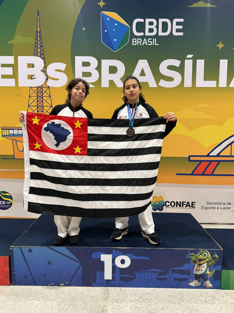 Duas atletas posam em um pódio segurando a bandeira do estado de São Paulo. Uma delas exibe uma medalha, diante de um painel de um evento esportivo escolar promovido pela CBDE Brasil.