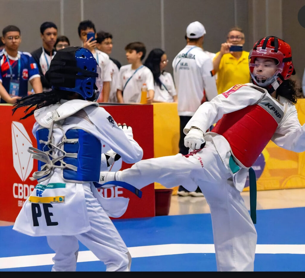 Duas atletas de taekwondo competem em uma luta, usando equipamentos de proteção nas cores azul e vermelho. Uma delas aplica um chute enquanto espectadores e outros participantes observam ao fundo.