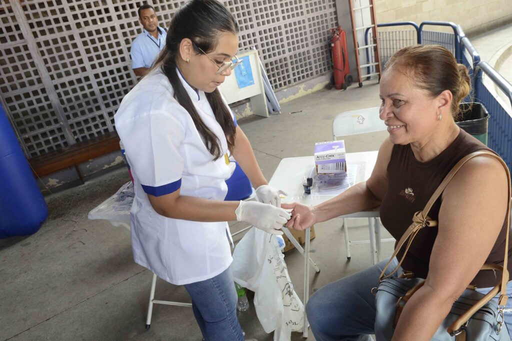 Profissional de saúde realizando um teste de glicemia em uma pessoa sentada durante uma ação de prevenção e cuidados com a saúde.