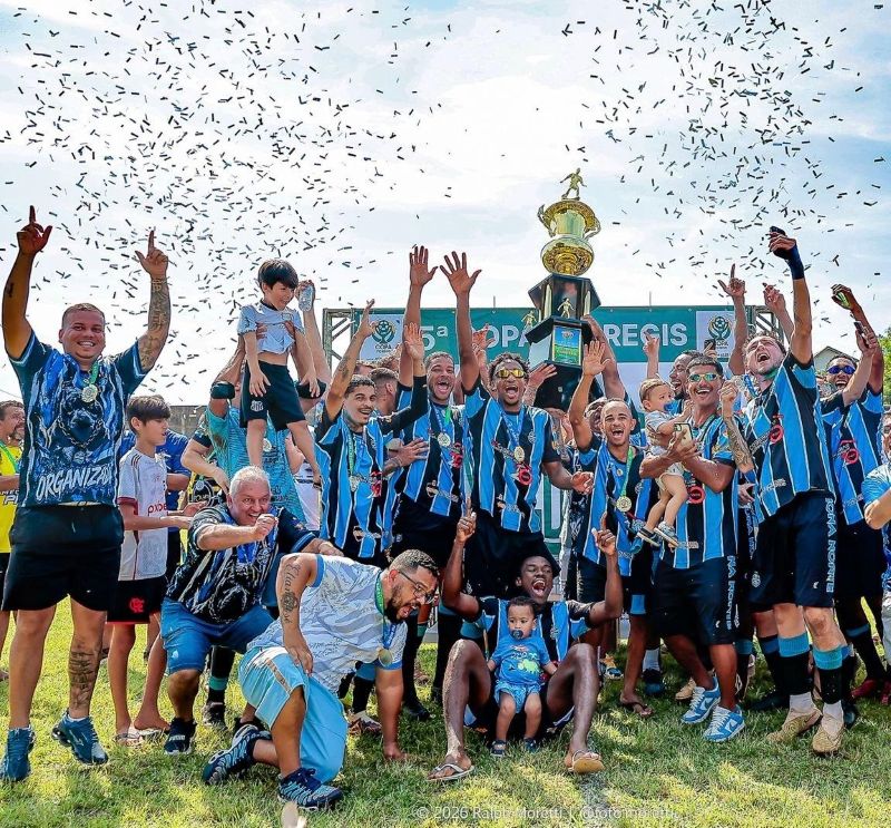 Um time de futebol comemora a vitória sob uma chuva de confetes com um troféu dourado ao fundo.
A imagem retrata um momento de exultação de um time de futebol masculino após uma vitória, com confetes caindo do céu e fitas azuis penduradas. No centro da imagem, um grande troféu dourado com uma estatueta de um atleta no topo é erguido por várias mãos. Os jogadores estão vestidos com uniformes de futebol nas cores azul e preto, com listras verticais, e muitos ostentam medalhas no pescoço. Alguns jogadores estão comemorando levantando os braços, tirando fotos com seus celulares ou exibindo o troféu. Um jogador carrega um bebê no colo, sorrindo para a câmera. Há outros adultos e crianças ao redor, alguns carregando bolsas e outros celebrando. A atmosfera é de pura alegria e triunfo. O gramado verde e o céu azul com algumas nuvens completam a cena, sugerindo um dia ensolarado e festivo. As cores predominantes são o azul, preto, dourado e verde, criando um contraste vibrante.