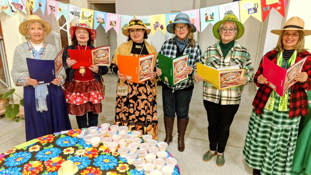 Grupo de seis pessoas vestidas com roupas e chapéus coloridos, segurando pastas ou cadernos. À frente há uma mesa decorada com toalha florida e vários recipientes plásticos. Ao fundo, bandeirinhas decorativas sugerem um evento cultural ou artístico.