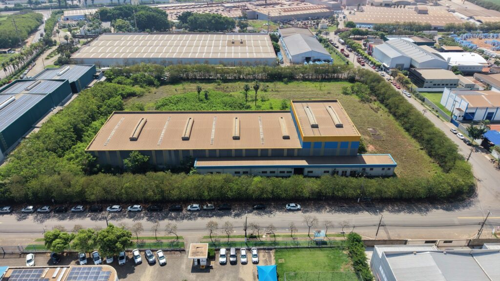 Vista aérea de galpão industrial edificado em distrito empresarial, cercado por áreas verdes e outras instalações industriais.