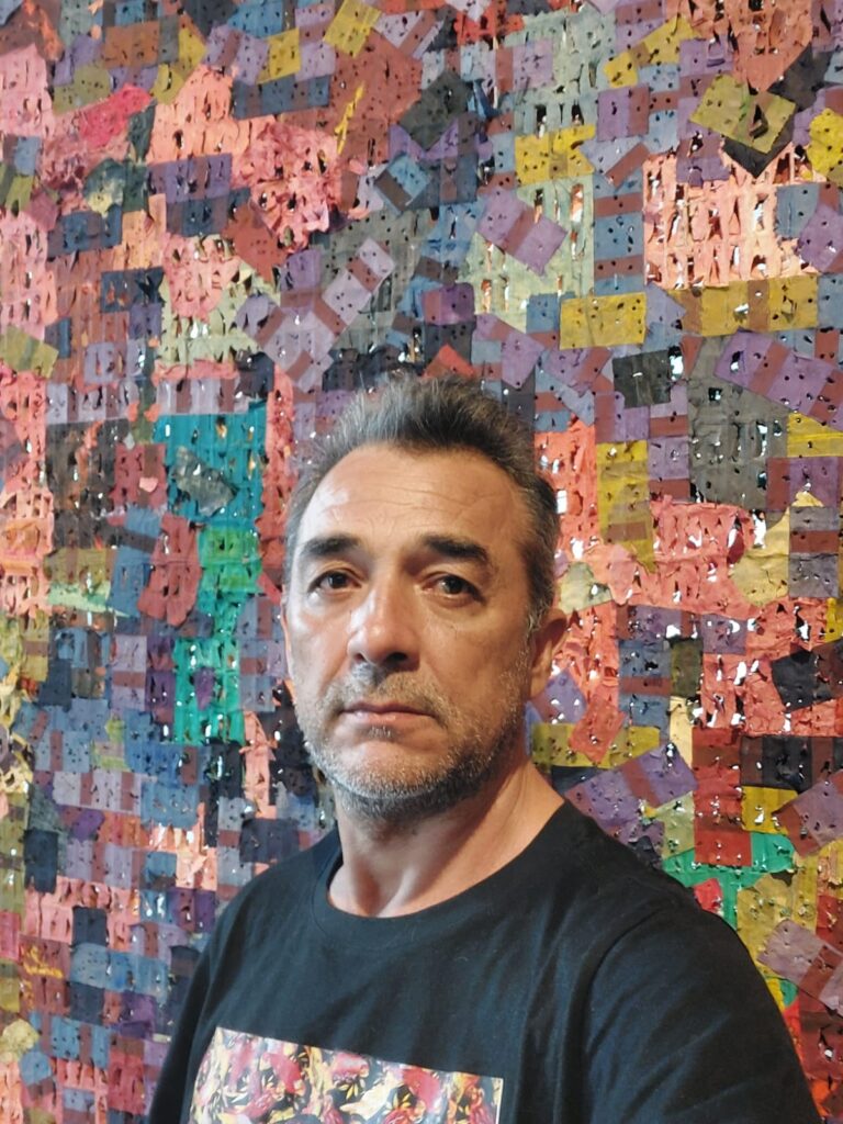 A imagem mostra uma pessoa em primeiro plano, usando uma camiseta preta, posicionada diante de um grande painel artístico formado por centenas de pequenos retalhos coloridos.