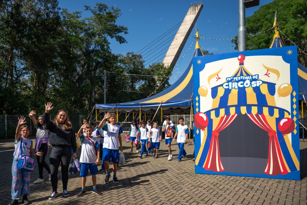 A imagem mostra um grupo de crianças e uma professora chegando animadamente ao Festival de Circo SP. Elas estão ao ar livre, em um dia ensolarado, e acenam sorridentes para a câmera. Ao fundo, vê-se uma grande tenda de circo azul e amarela e uma placa colorida com o nome do evento — “19º Festival de Circo SP”. O cenário transmite alegria e expectativa, típico de uma atividade cultural escolar.