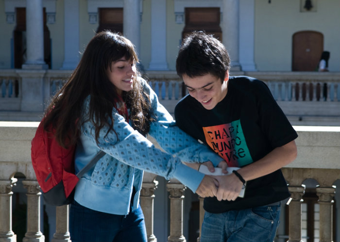 A imagem mostra duas pessoas jovens em um espaço aberto diante de um prédio com colunas. Uma delas usa mochila vermelha e casaco azul, enquanto a outra veste camiseta preta e jeans.