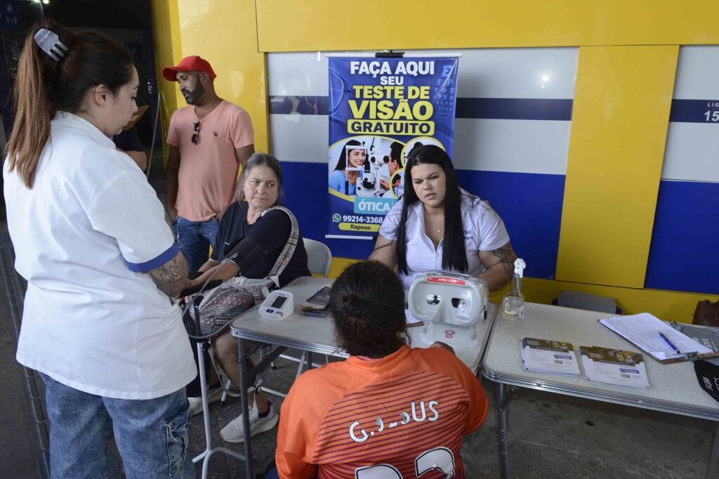 Atendimento de saúde comunitária com realização de teste de visão gratuito e aferição de pressão arterial em uma mesa de serviços.