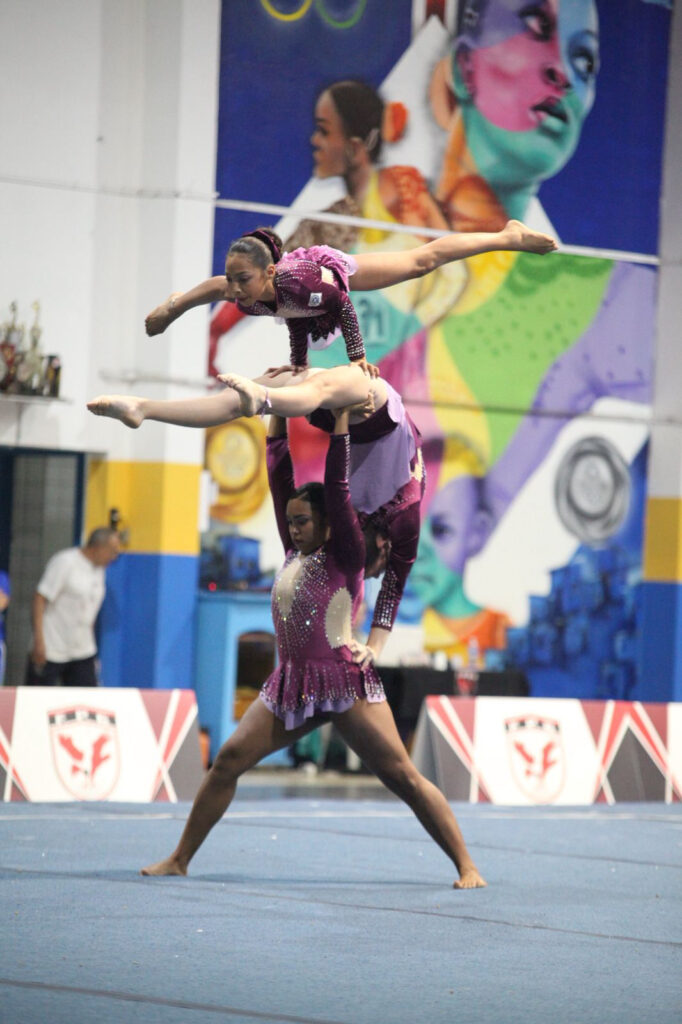 A imagem mostra um grupo de atletas realizando uma apresentação de ginástica acrobática em uma competição. Três participantes formam uma estrutura elevada, com uma pessoa sustentando outra acima da cabeça enquanto uma terceira executa uma posição de equilíbrio no topo.