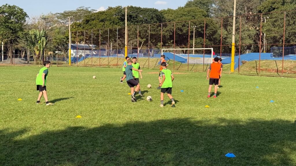 A imagem mostra um grupo de crianças e adolescentes treinando futebol em um campo gramado. Alguns usam coletes verdes e outros, coletes laranjas; há várias bolas e cones coloridos espalhados pelo campo. Ao fundo, vê-se uma quadra esportiva cercada por telas, com traves, árvores e postes. O dia está ensolarado, e uma grande sombra cobre a parte inferior do gramado.