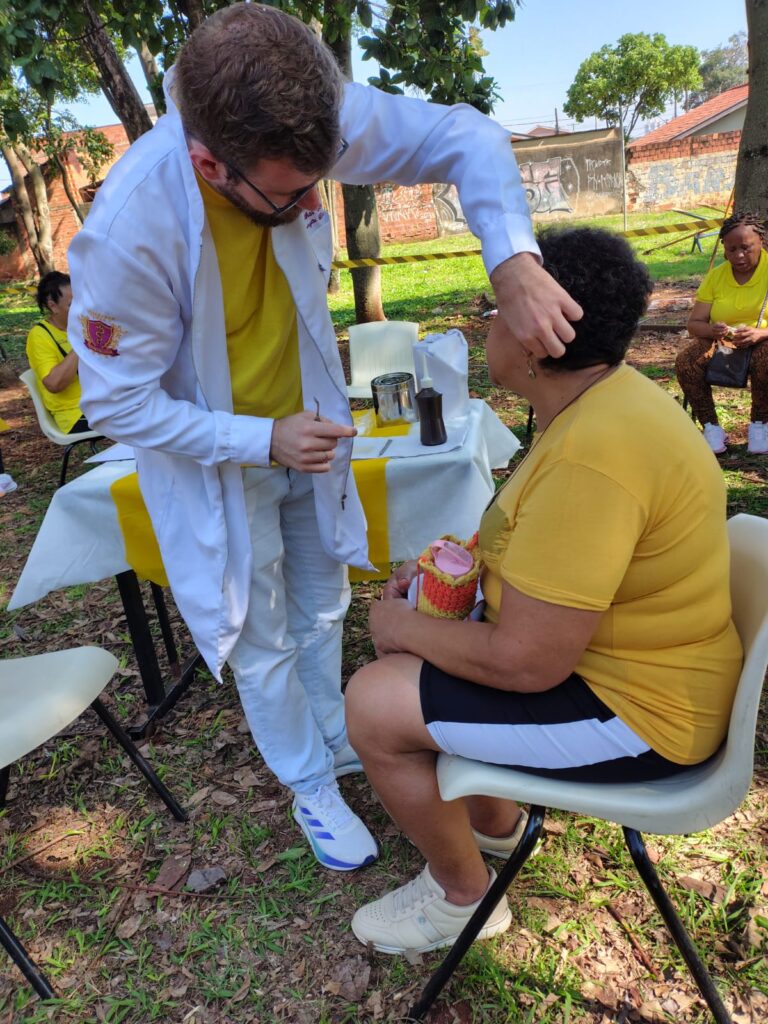Atendimento de auriculoterapia ao ar livre, com um profissional realizando o procedimento em uma participante sentada durante uma atividade comunitária.