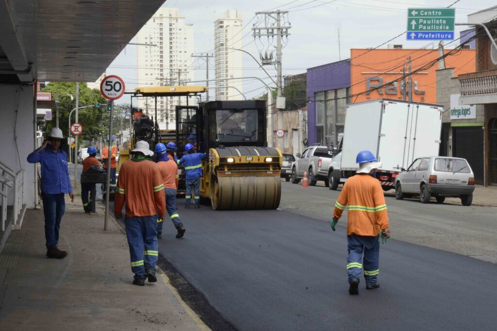 Equipe de trabalhadores realiza recapeamento asfáltico em uma avenida, utilizando máquinas de pavimentação e compactação enquanto o trânsito segue parcialmente liberado.