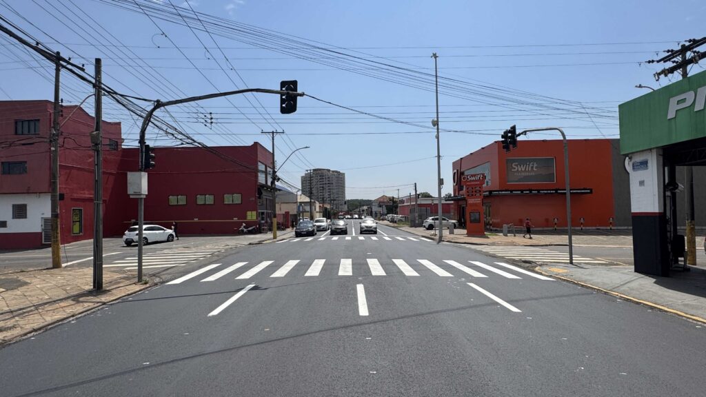 Cruzamento urbano com sinalização viária renovada, faixa de pedestres e pavimento recém-recapeado em uma área comercial.