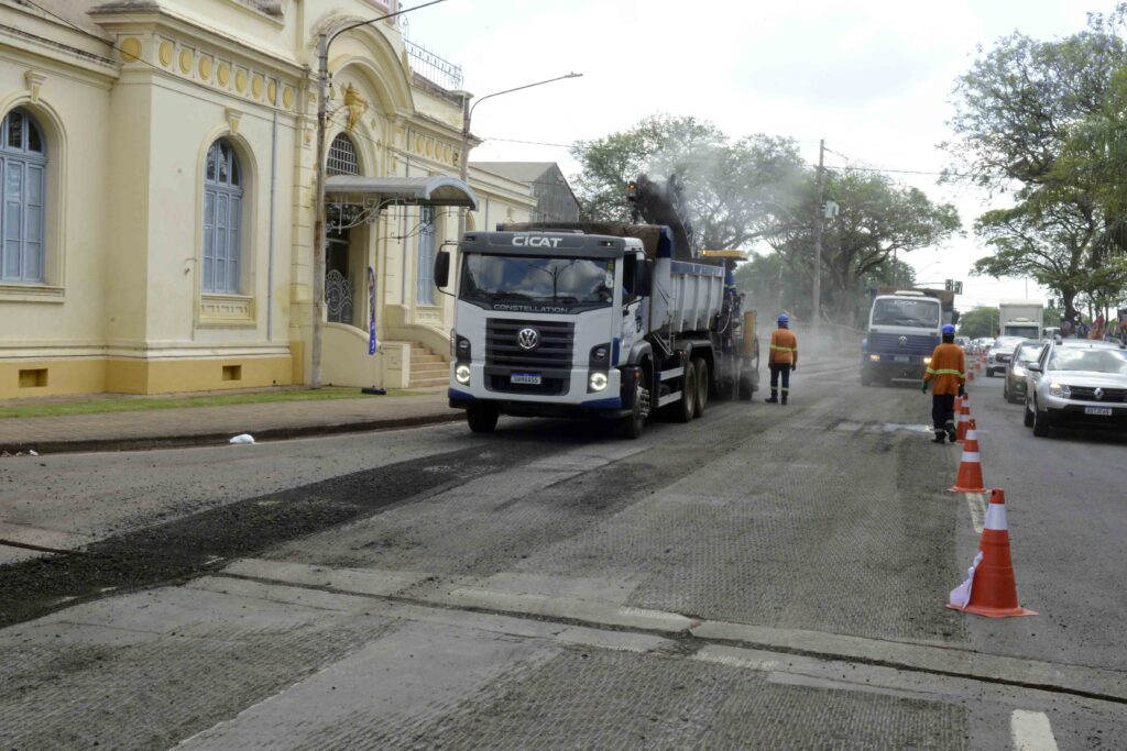 Caminhão e operários executam obras de recuperação do pavimento em via urbana sinalizada com cones, próximo a prédios históricos e áreas arborizadas.